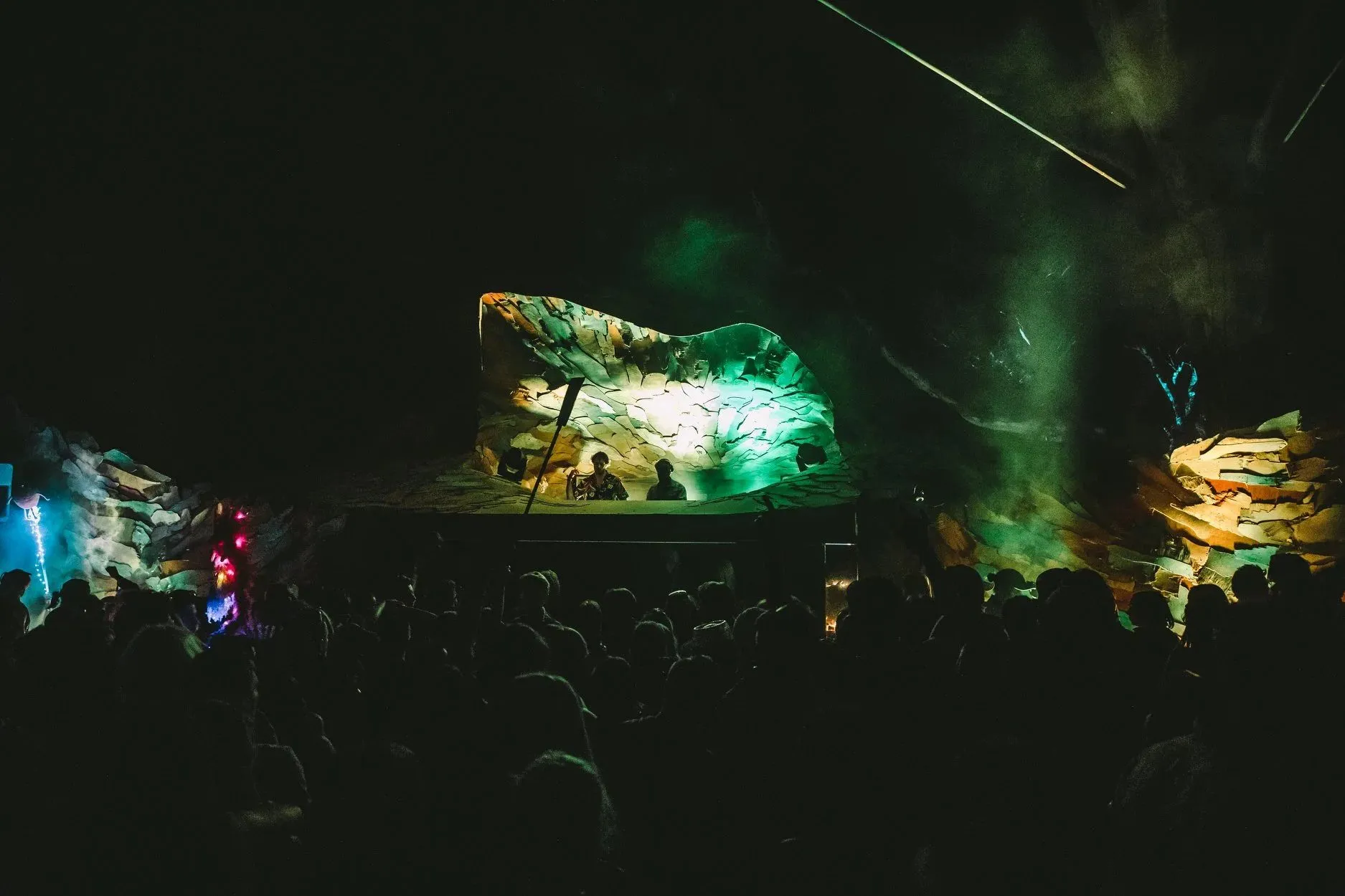 Sweeping bark-clad winged stage form glowing green and gold above the DJ booth, enveloping the night-time crowd