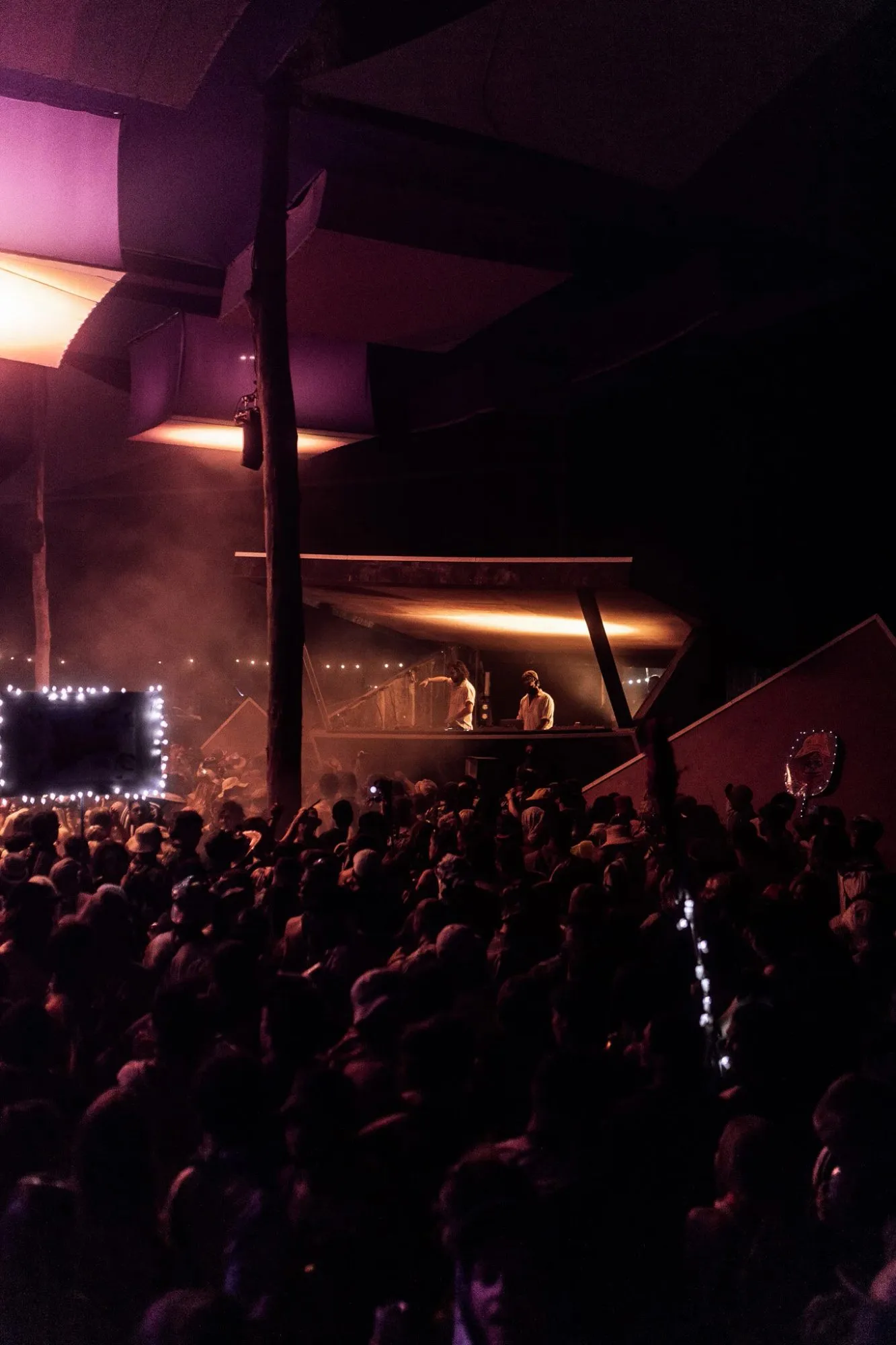 DJs in an angular timber booth beneath a cantilevered roof, amber light and purple-lit cubic canopies above the night crowd