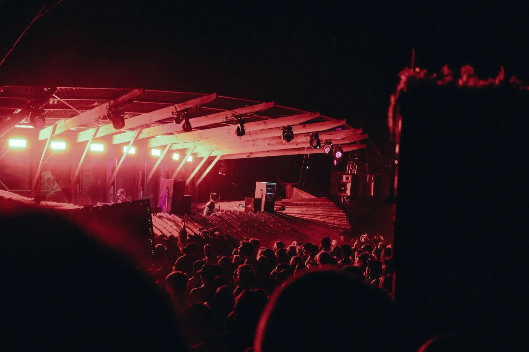 Cantilevered timber stage roof washed in red light over the DJ booth, with a bamboo facade and night crowd below