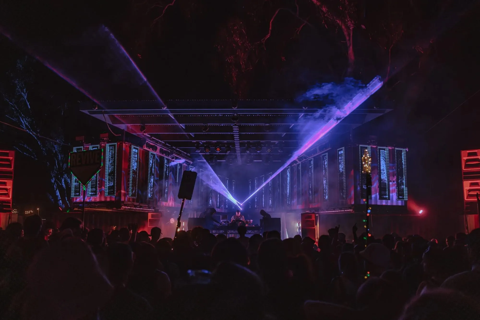 Night stage flanked by two container wings lit with forest projections, blue lasers crossing above the DJ and crowd