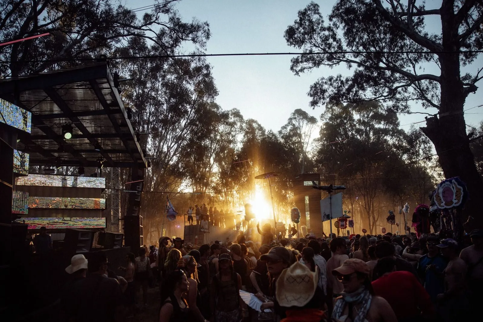 Golden sunset breaking through gum trees behind the Deep Jungle stage, LED screens and shade poles over the festival crowd
