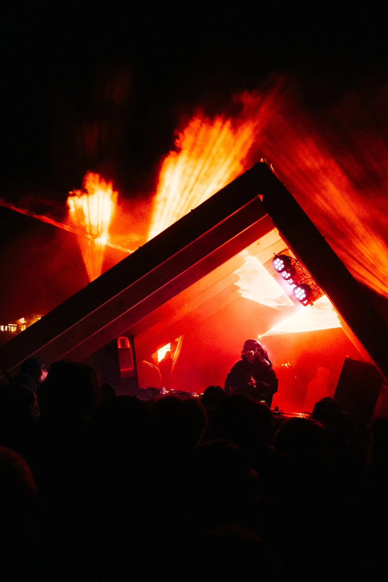 DJ framed inside the offset triangular timber stage structure, backlit by red beams and haze at night