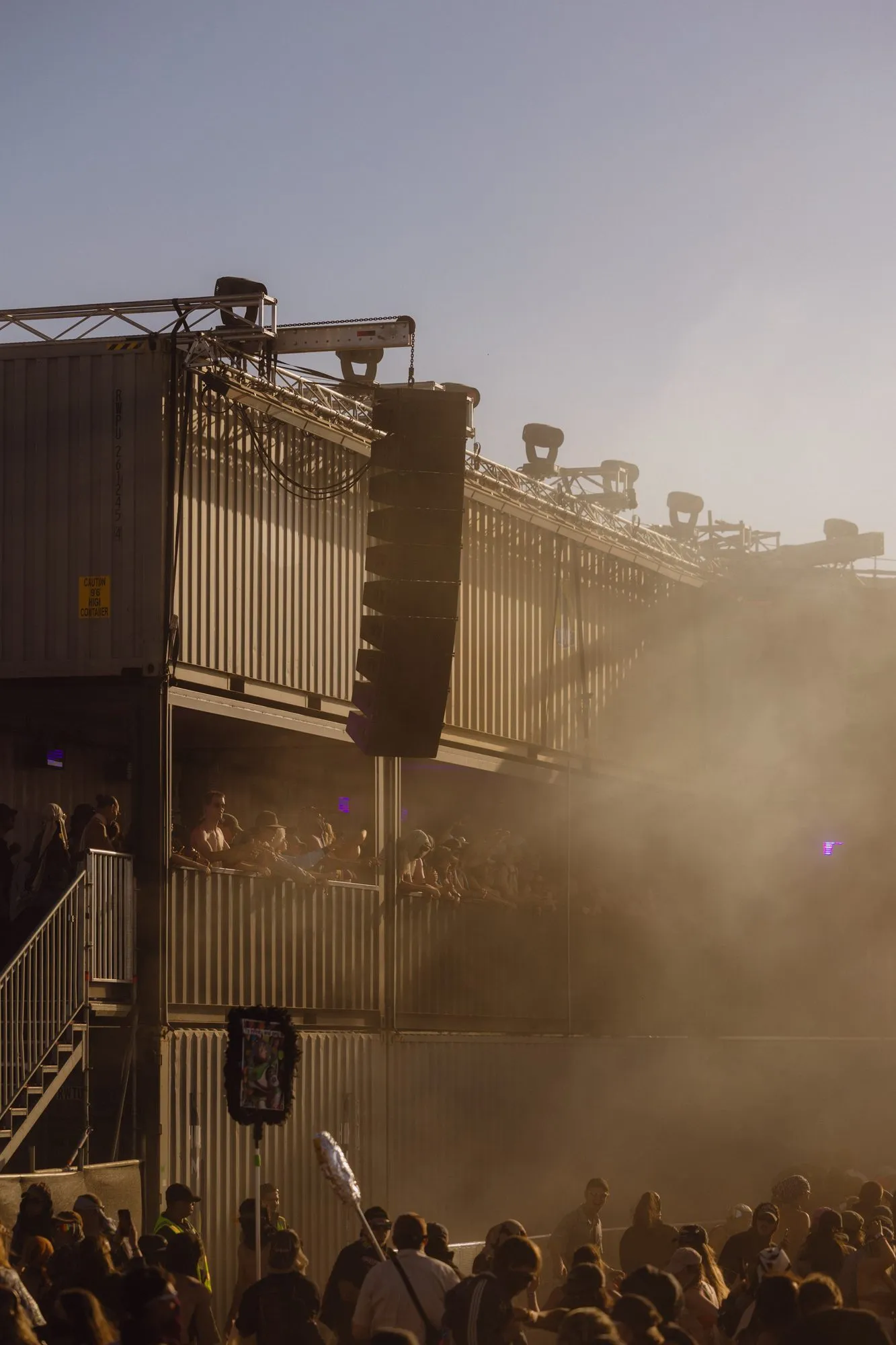 Three tiers of stacked shipping containers in golden dusty haze, festivalgoers watching from the container balconies above the crowd