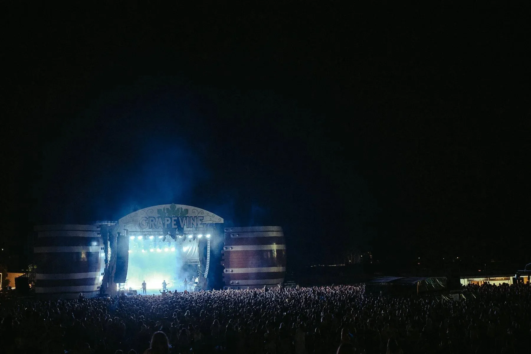 Festival stage framed by two giant striped wine-barrel forms beneath an arched Grapevine sign, the night crowd stretching into the dark