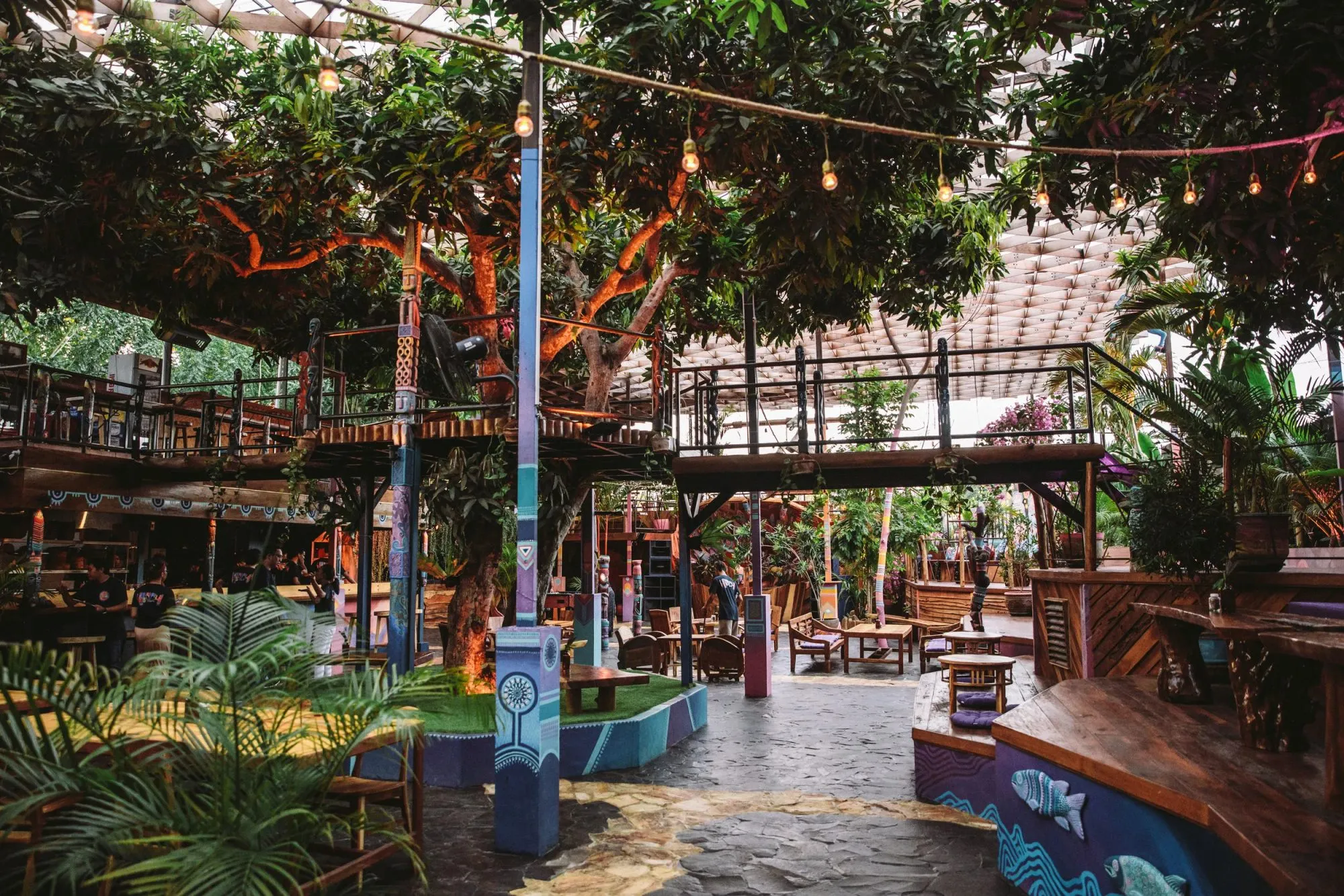 Venue courtyard beneath mango trees and a timber lattice canopy, with raised walkways, hand-painted columns and festoon lighting