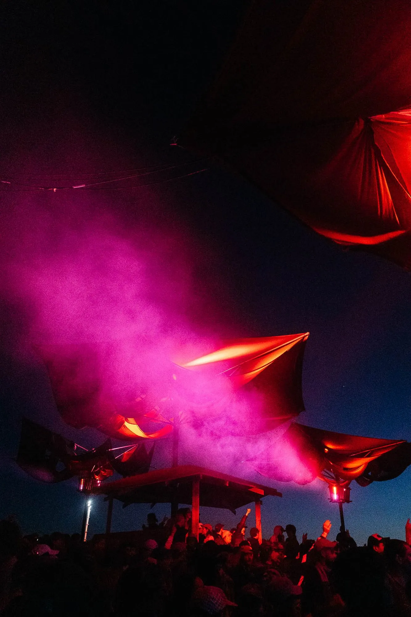 Fabric canopies washed in red and pink light above the stage structure at dusk, haze drifting into the night sky