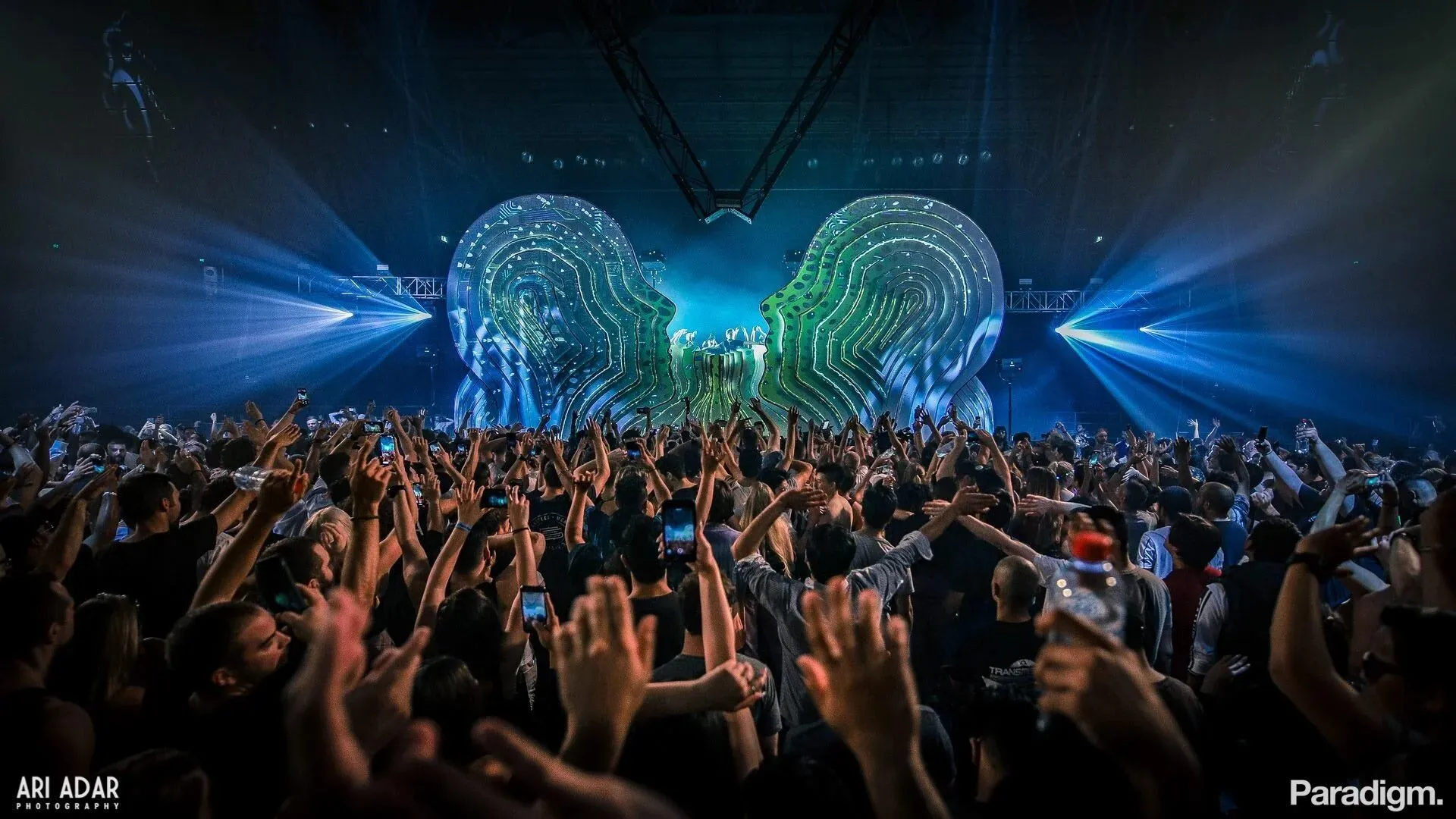 Two towering layered face sculptures meeting above the DJ across the stage, lit green and blue over an arena crowd