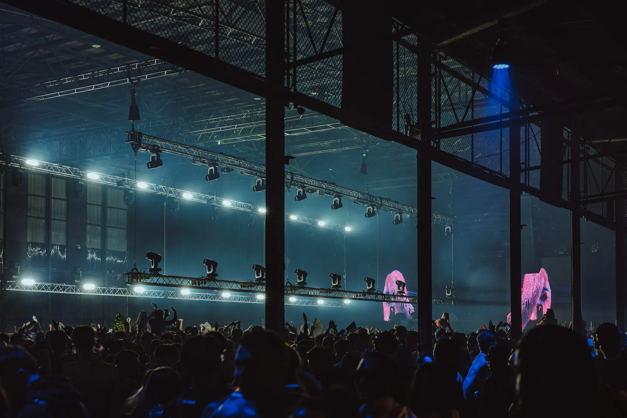 Vast industrial hall with steel trusses and lighting rigs over a dense crowd, pink LED figures glowing at the stage