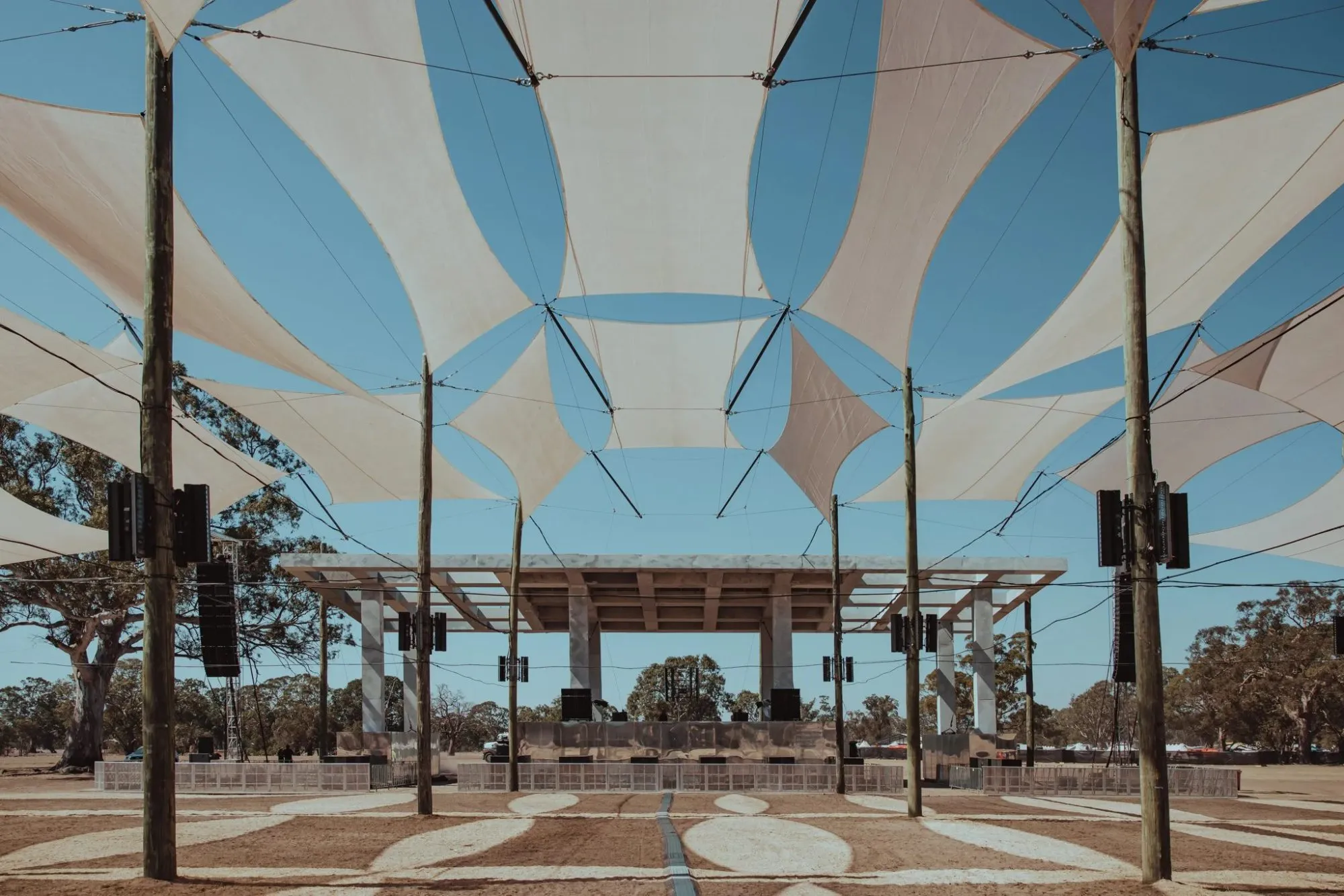 White shade sails stretched between timber poles over a patterned arena, leading to Pitch One's gridded concrete roof canopy under a blue sky