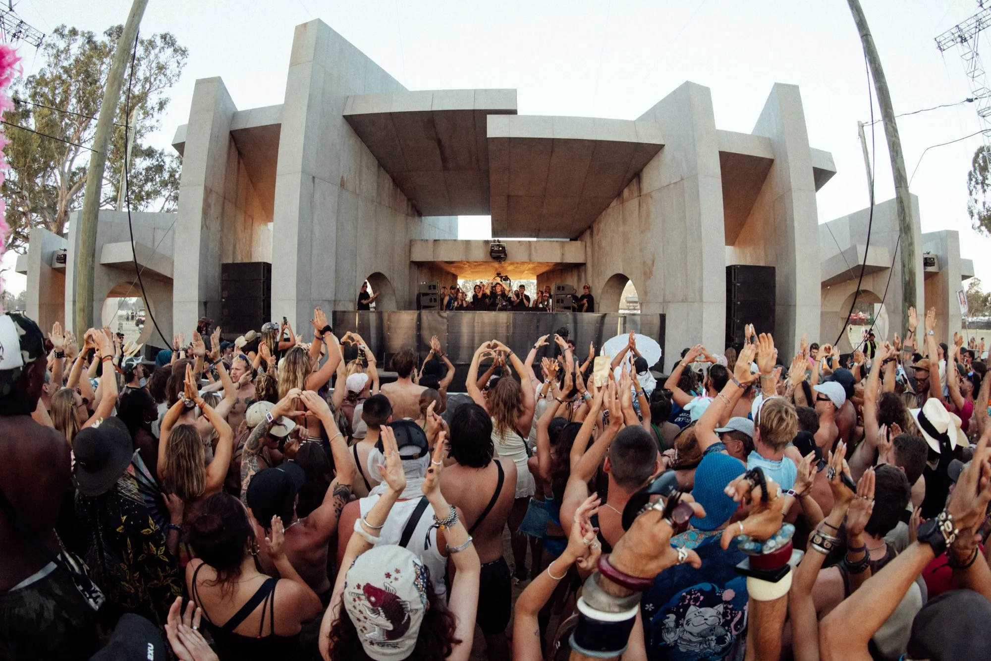 Dense daytime crowd with arms raised facing Pitch One's full brutalist concrete stage head-on, DJs at the booth beneath the central arch