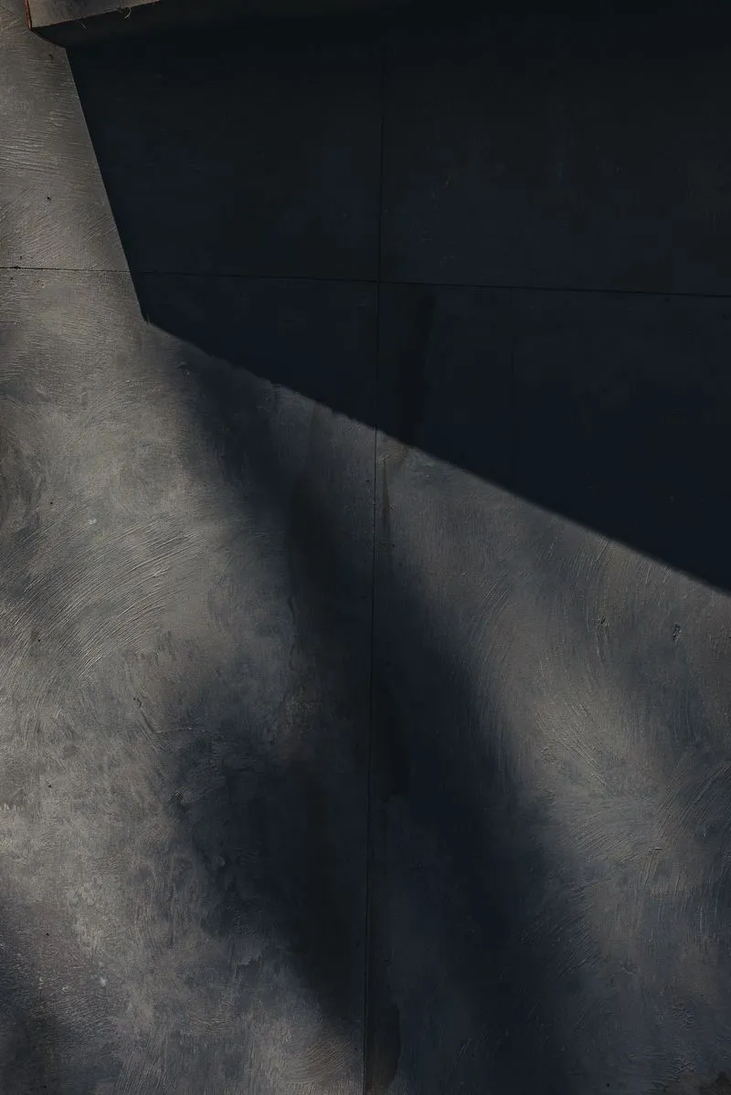 Raw board-formed concrete face of the Pitch One stage, a hard diagonal of sunlight cutting across the textured grey surface into deep shadow