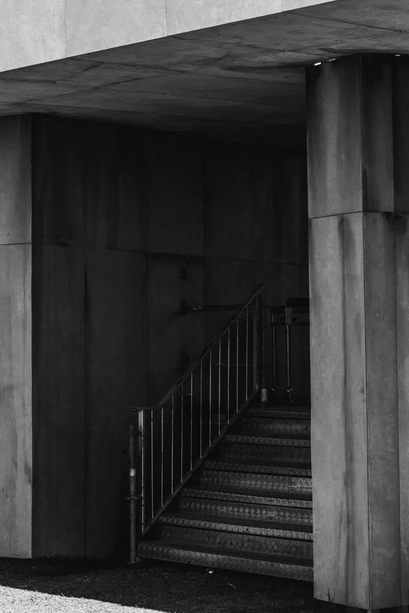 Metal checkerplate stairway with a black handrail rising into the shadowed underside of the Pitch One stage, framed by raw board-formed concrete walls