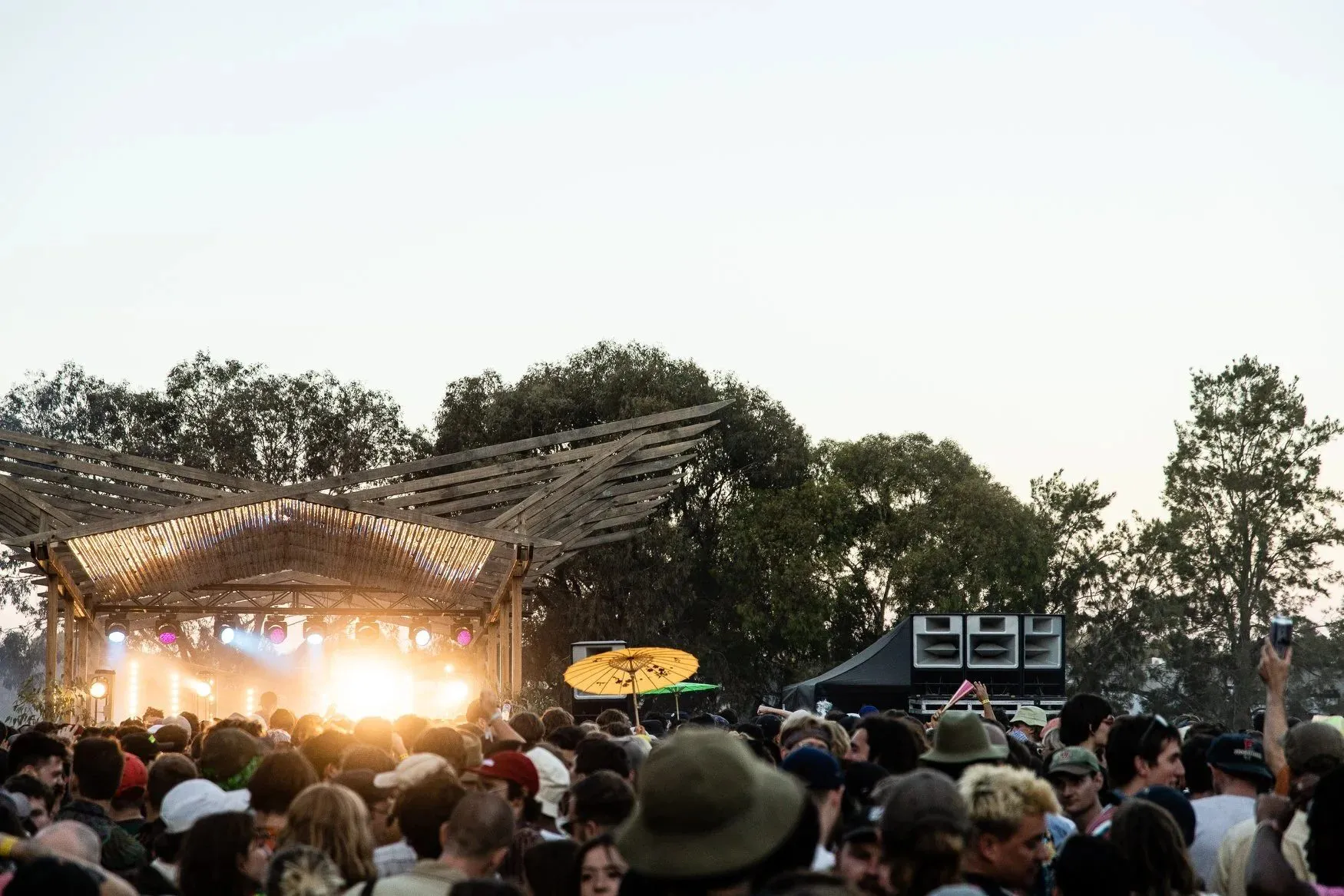 Timber pergola with splayed wing-like rafters over a glowing stage at dusk, paper parasols raised among the crowd