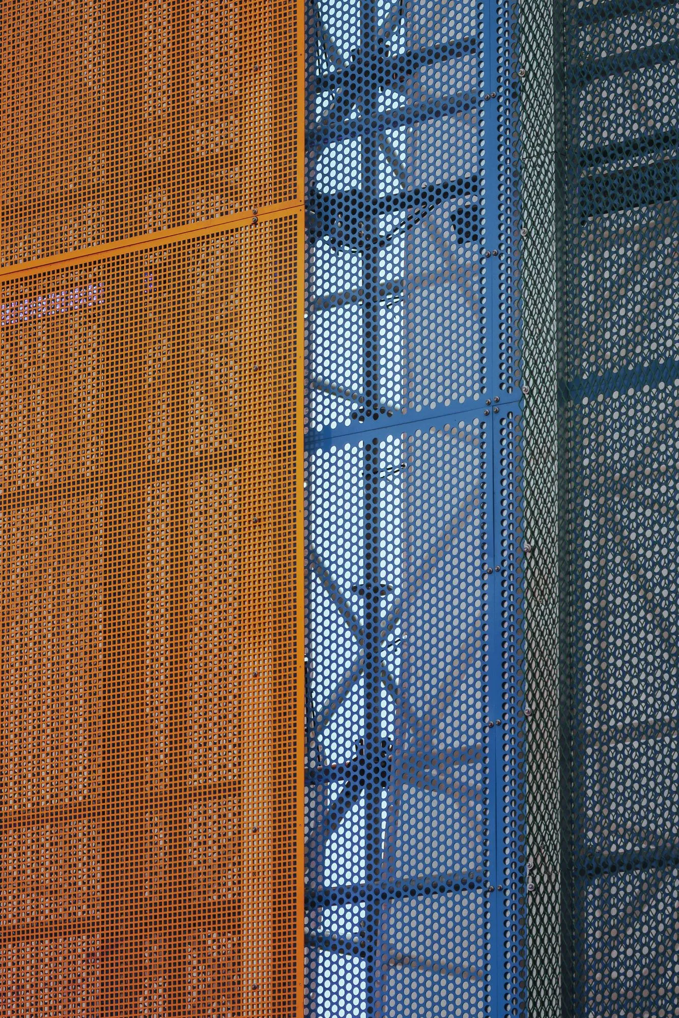 Detail of layered perforated metal screens in orange, blue and green, steel bracing showing through the mesh