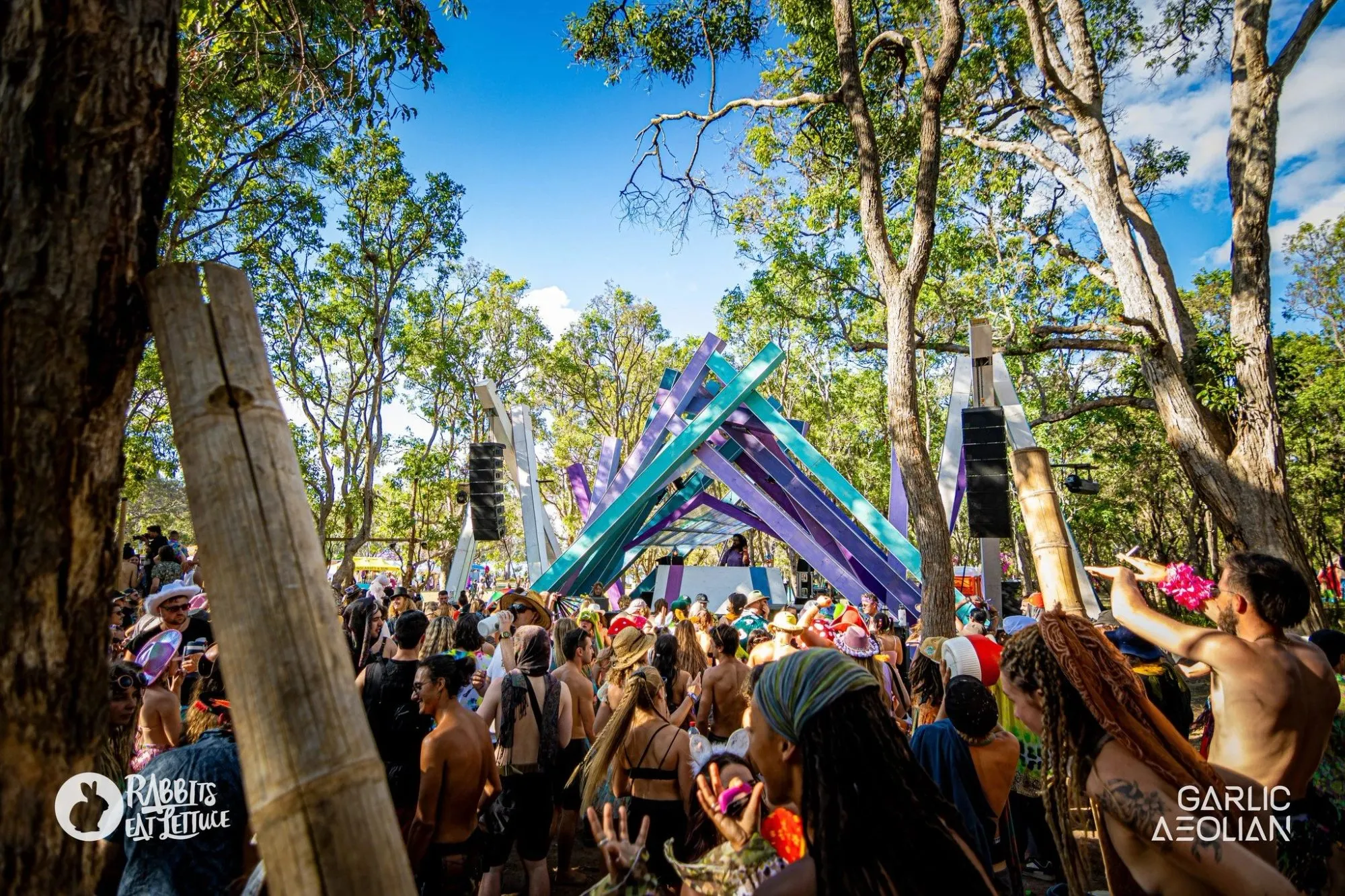 Purple and teal beams interlocking into a sculptural peak over the stage, crowd dancing beneath gum trees on a sunny day