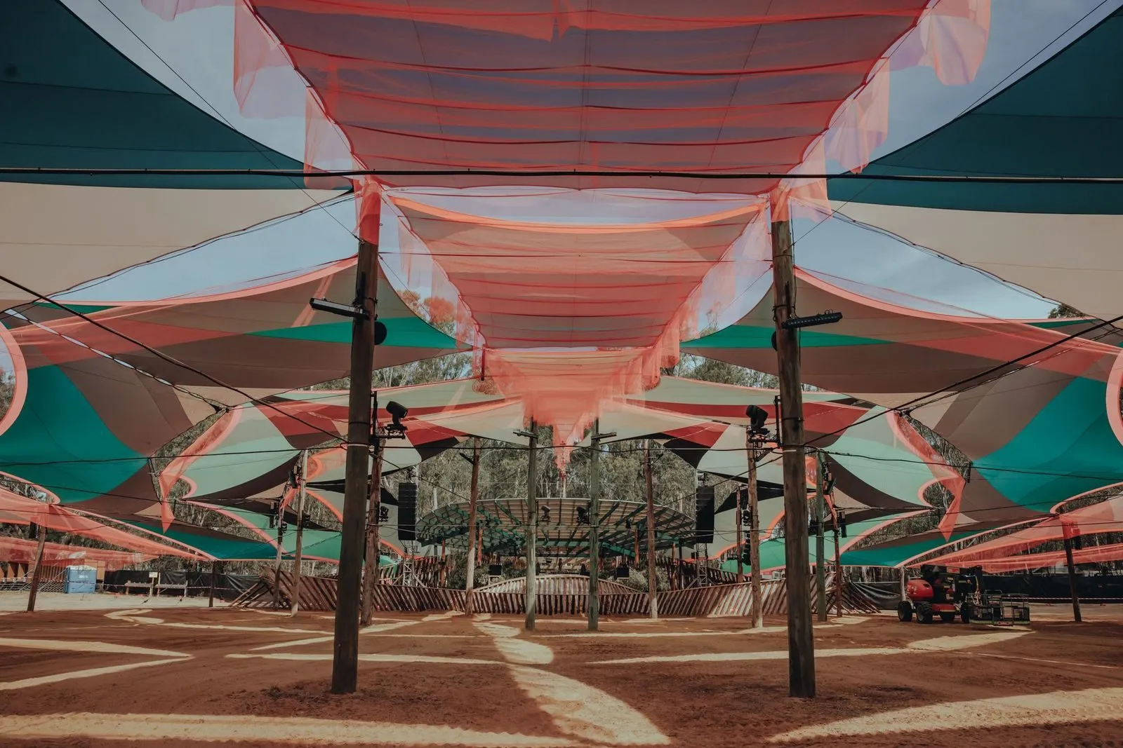 Layers of pink, teal and cream shade sails on timber poles, looking toward the circular timber-slat stage enclosure